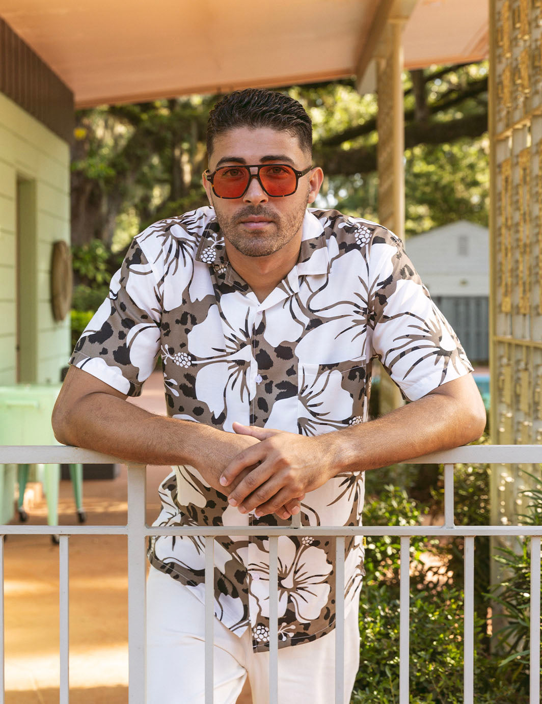 Man wearing a floral shirt and sunglasses leaning on a railing outdoors.