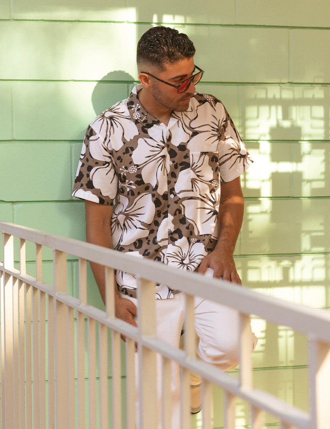 Man wearing a floral shirt leaning against a light green wall.