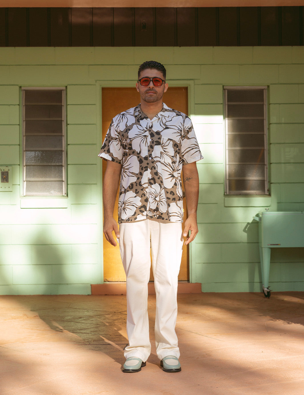 Man wearing a floral shirt and white pants standing in front of a green building.